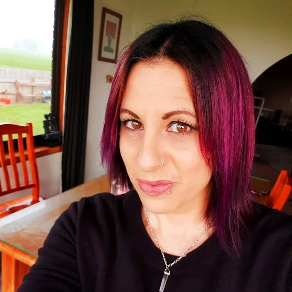 [Image Description: A selfie photo of Tess, an Indigenous woman with pale skin and short straight pink hair with brown roots. She is wearing a black top,  silver necklace and a silver nose ring.]