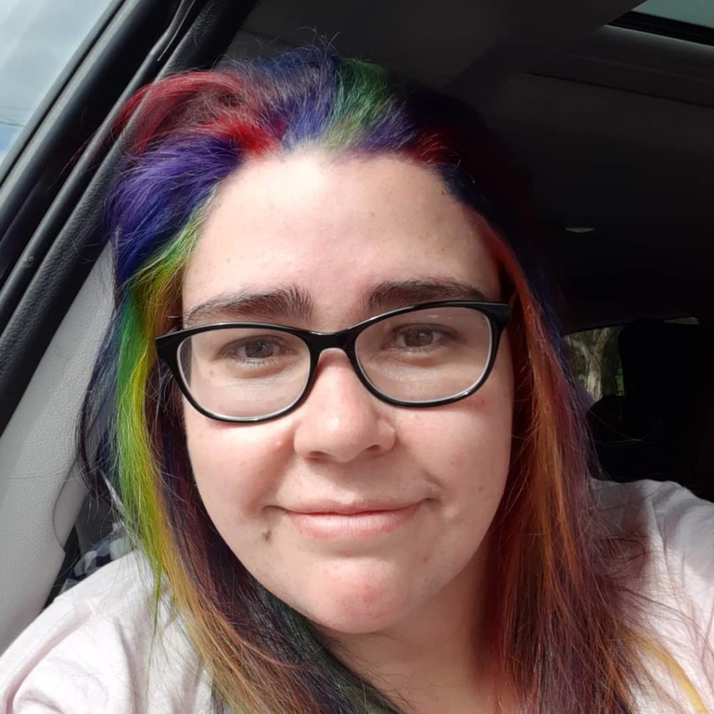 [Image Description: A selfie of Renay, an Indigenous woman wearing a pink tshirt, and black rimmed glasses. She has rainbow coloured shoulder length hair.]